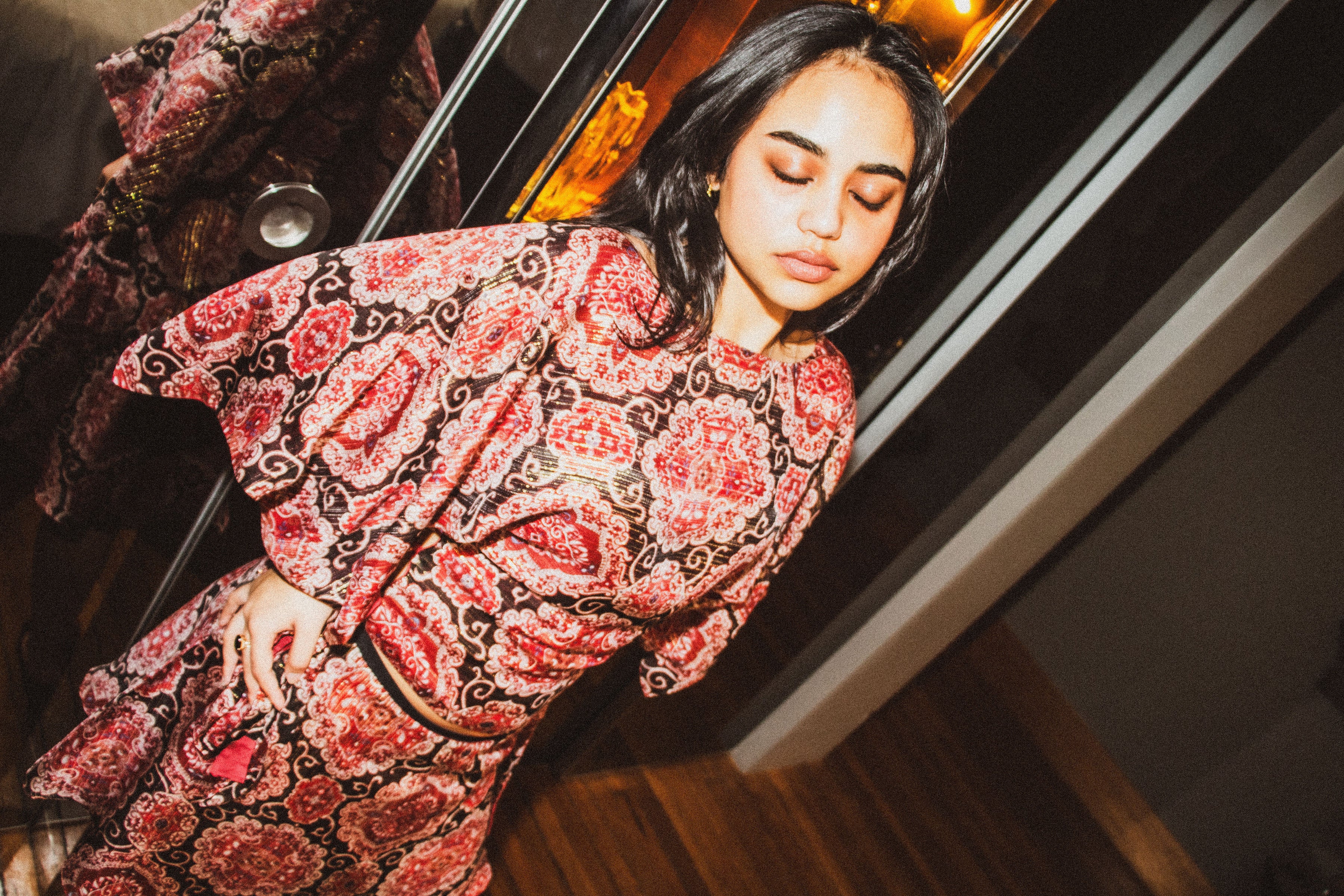 A woman wearing a red and black paisley patterned long-sleeve dress, leaning against a glass door in an indoor setting.