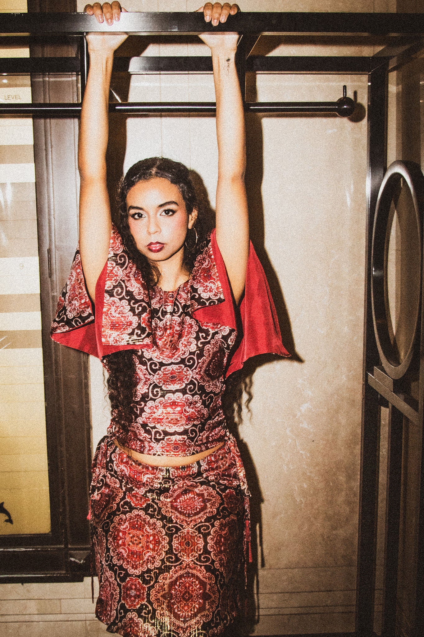 A woman hangs from a black metal frame in a doorway, wearing a red and black patterned two-piece outfit.