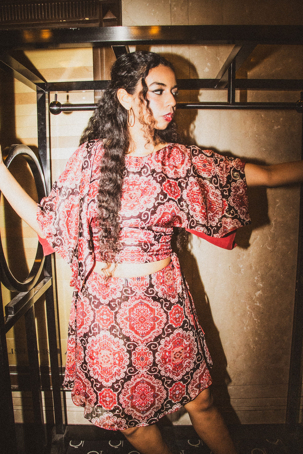 A woman modeling a red and black patterned two-piece set consisting of a short-sleeve crop top and a matching flared skirt, posed in front of a textured wall.