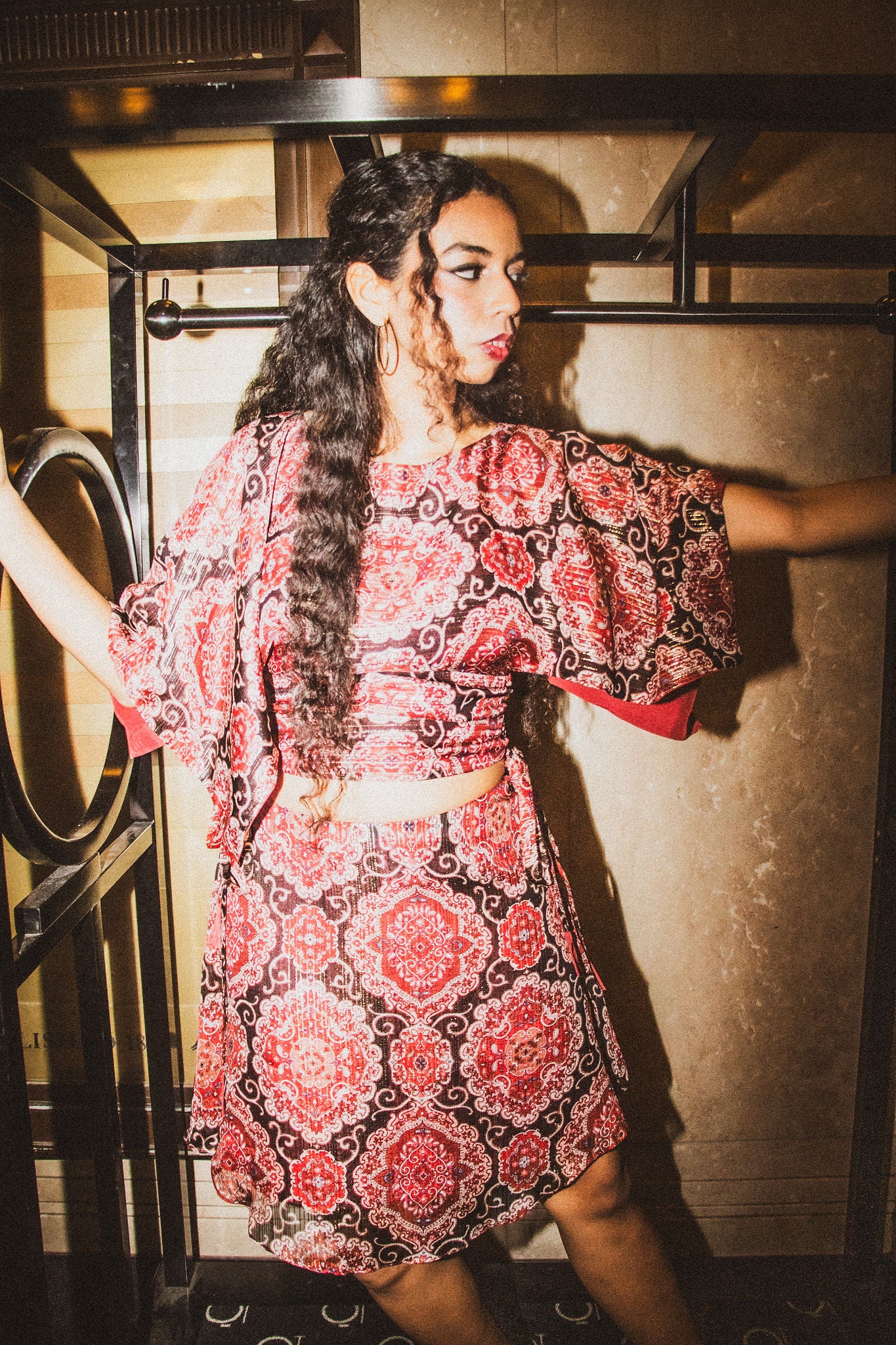 A woman modeling a red and black patterned two-piece set consisting of a short-sleeve crop top and a matching flared skirt, posed in front of a textured wall.