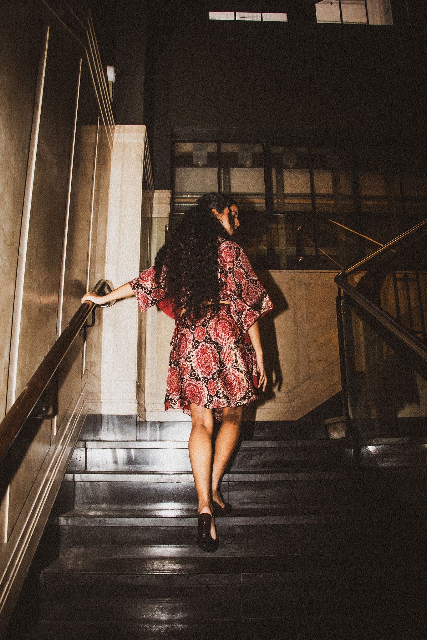 A woman with long curly dark hair walks up a dark staircase wearing a short red and pink floral print dress and black heels, holding onto a metal railing in a dimly lit interior.