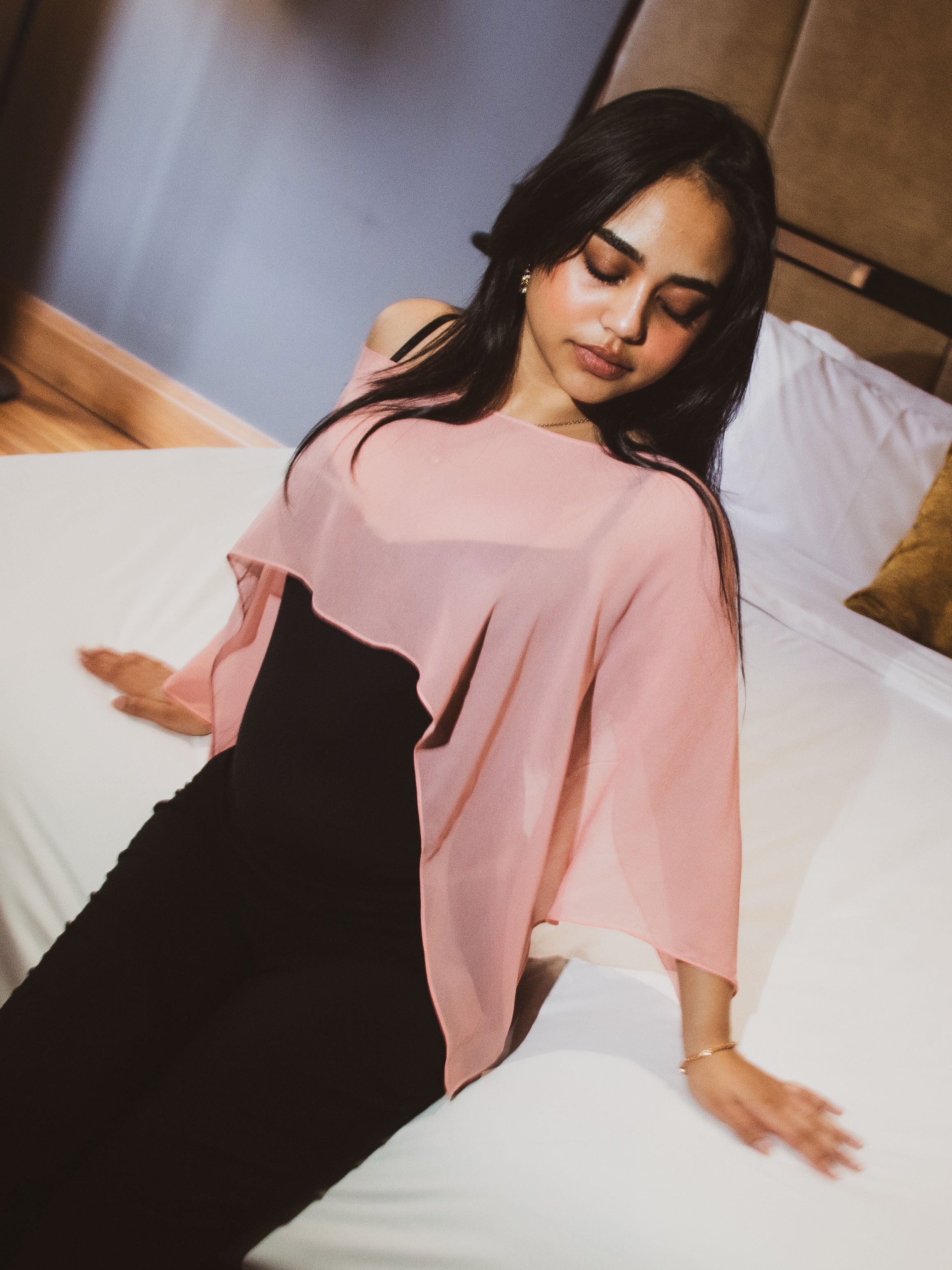 A woman reclining on a white bed wearing a sheer light pink draped top over a black dress, with a wooden headboard and pillows in the background.
