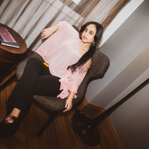 A woman reclining in a gray armchair in a dimly lit room, wearing a light pink blouse, black pants, and black sandals.