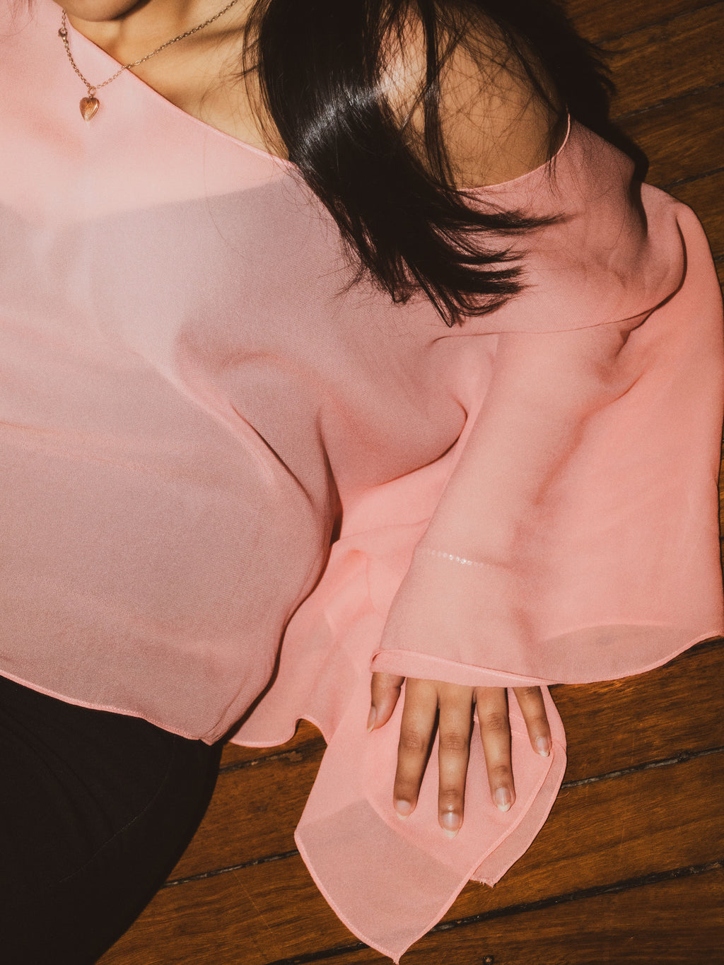 A close-up of a person wearing a sheer, light pink off-the-shoulder blouse, resting on a dark wooden floor.