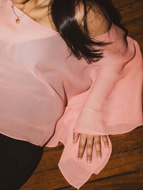 A close-up of a person wearing a sheer, light pink off-the-shoulder blouse, resting on a dark wooden floor.