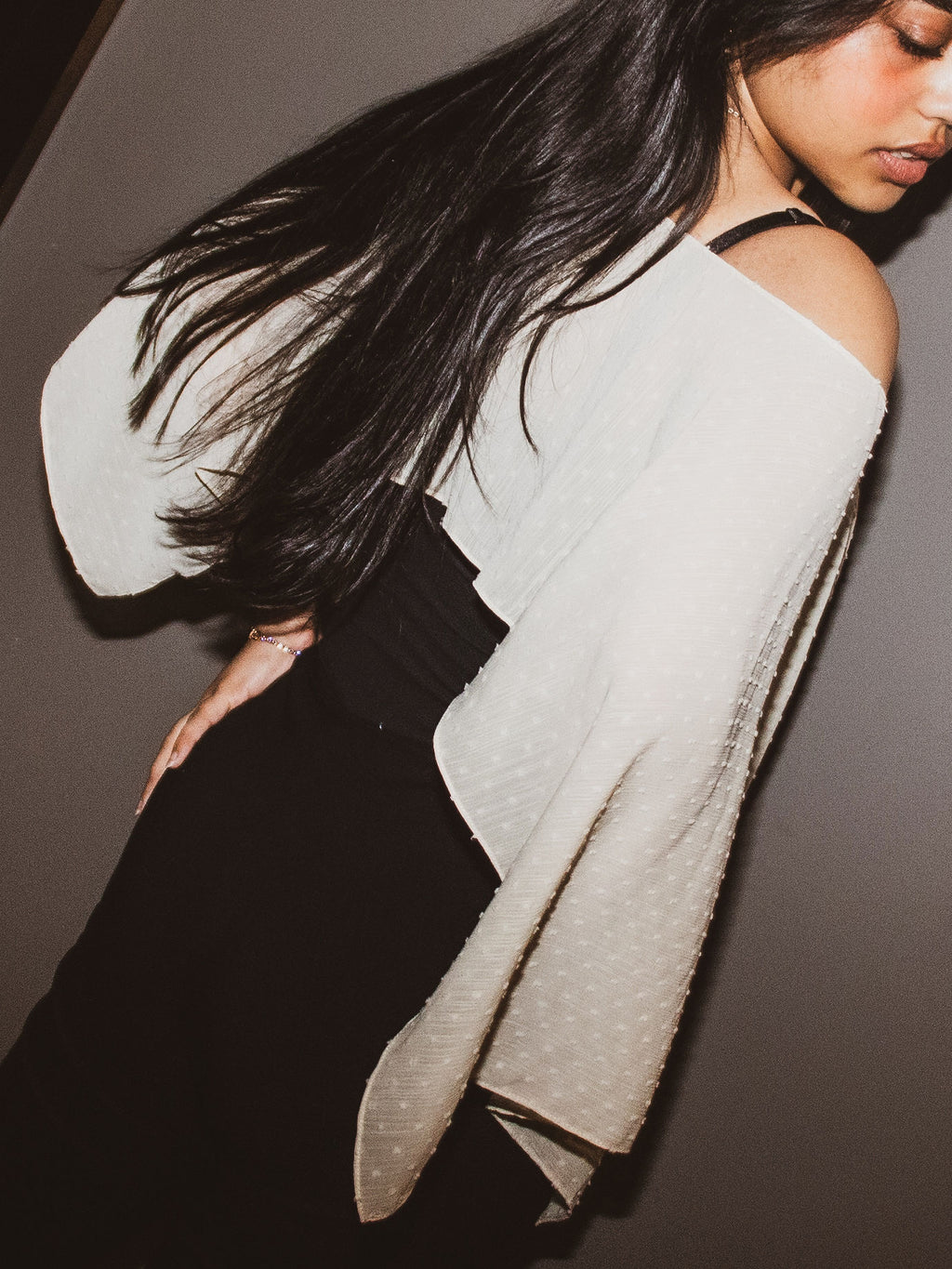 A woman wearing a textured off-white knit poncho-style top with wide draped sleeves and black bottoms, photographed against a dark gray background.