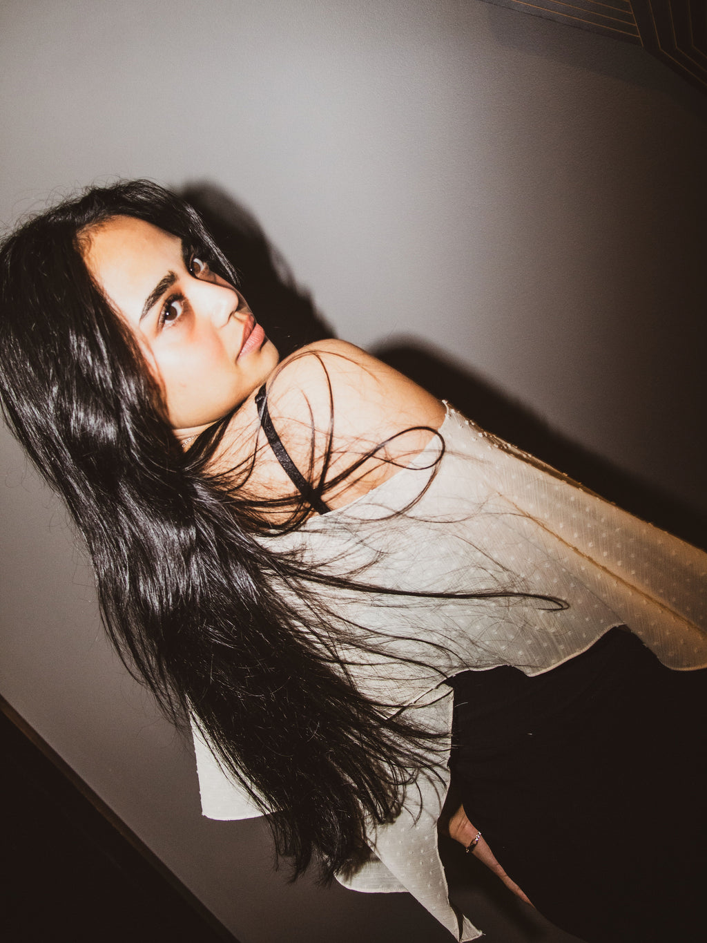 A woman with long, dark hair wearing a sheer, off-the-shoulder white top with subtle polka dots, posed against a dark gray wall.