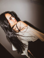 A woman with long, dark hair wearing a sheer, off-the-shoulder white top with subtle polka dots, posed against a dark gray wall.