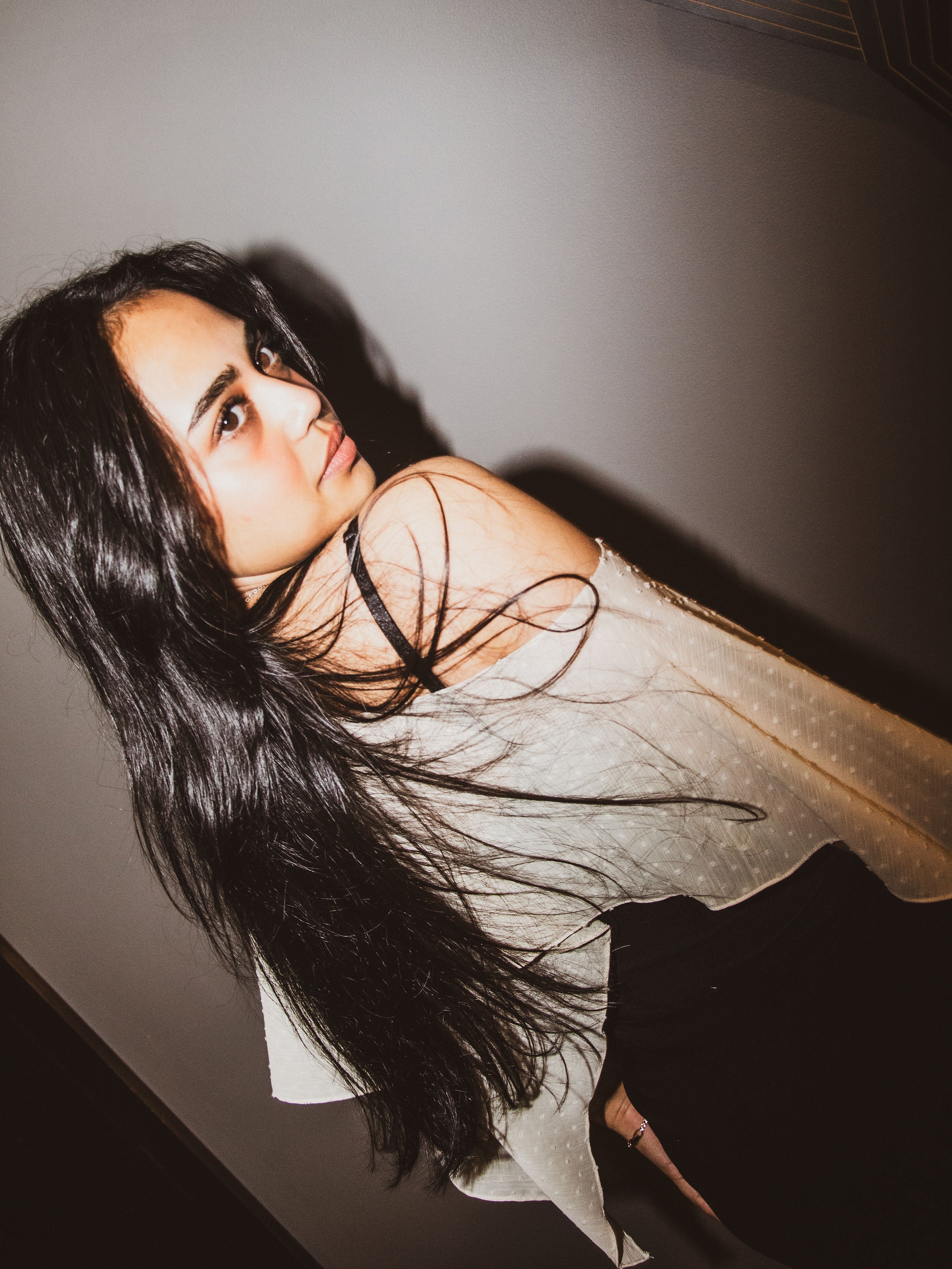 A woman with long, dark hair wearing a sheer, off-the-shoulder white top with subtle polka dots, posed against a dark gray wall.