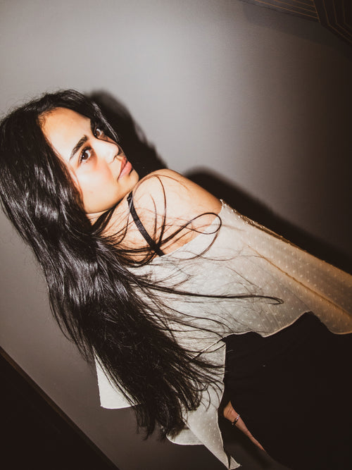 A woman with long, dark hair wearing a sheer, off-the-shoulder white top with subtle polka dots, posed against a dark gray wall.