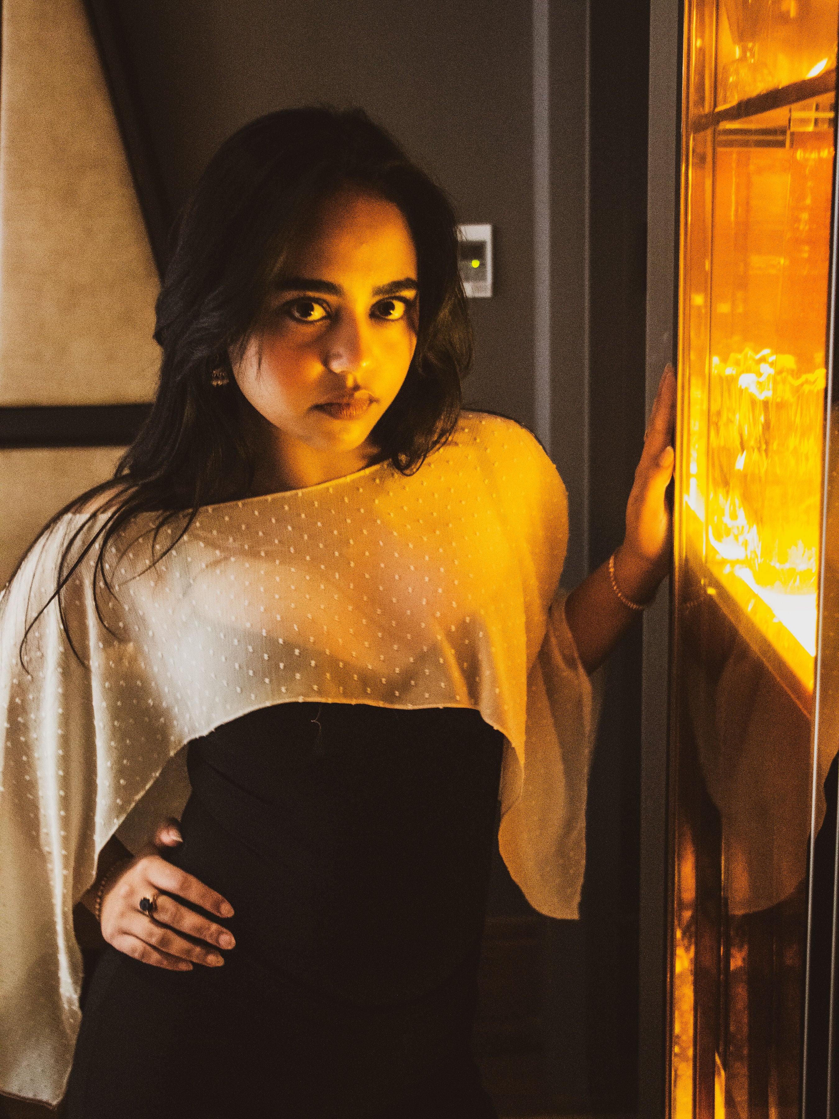 A woman wearing a sheer white dotted cape-style top over a black dress, posing next to a brightly lit warm-toned glass display case in an indoor setting.