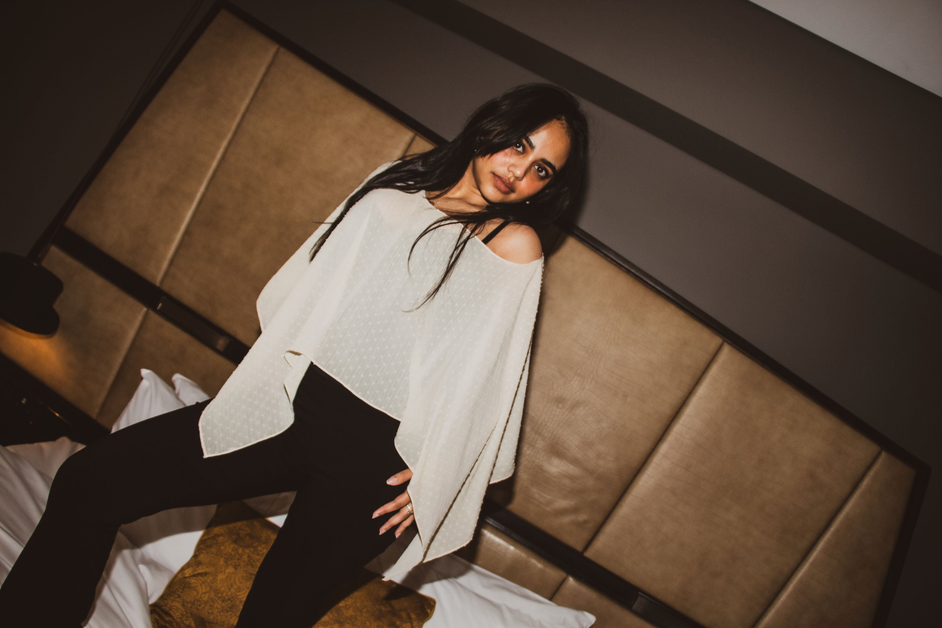 A woman with long dark hair stands on a bed wearing a white off-the-shoulder top and black pants, posed against a brown padded headboard in a dimly lit bedroom.