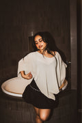 A woman posing in a bathroom next to a bathtub, wearing a loose, oversized light beige knit top and a black skirt with silver studs.