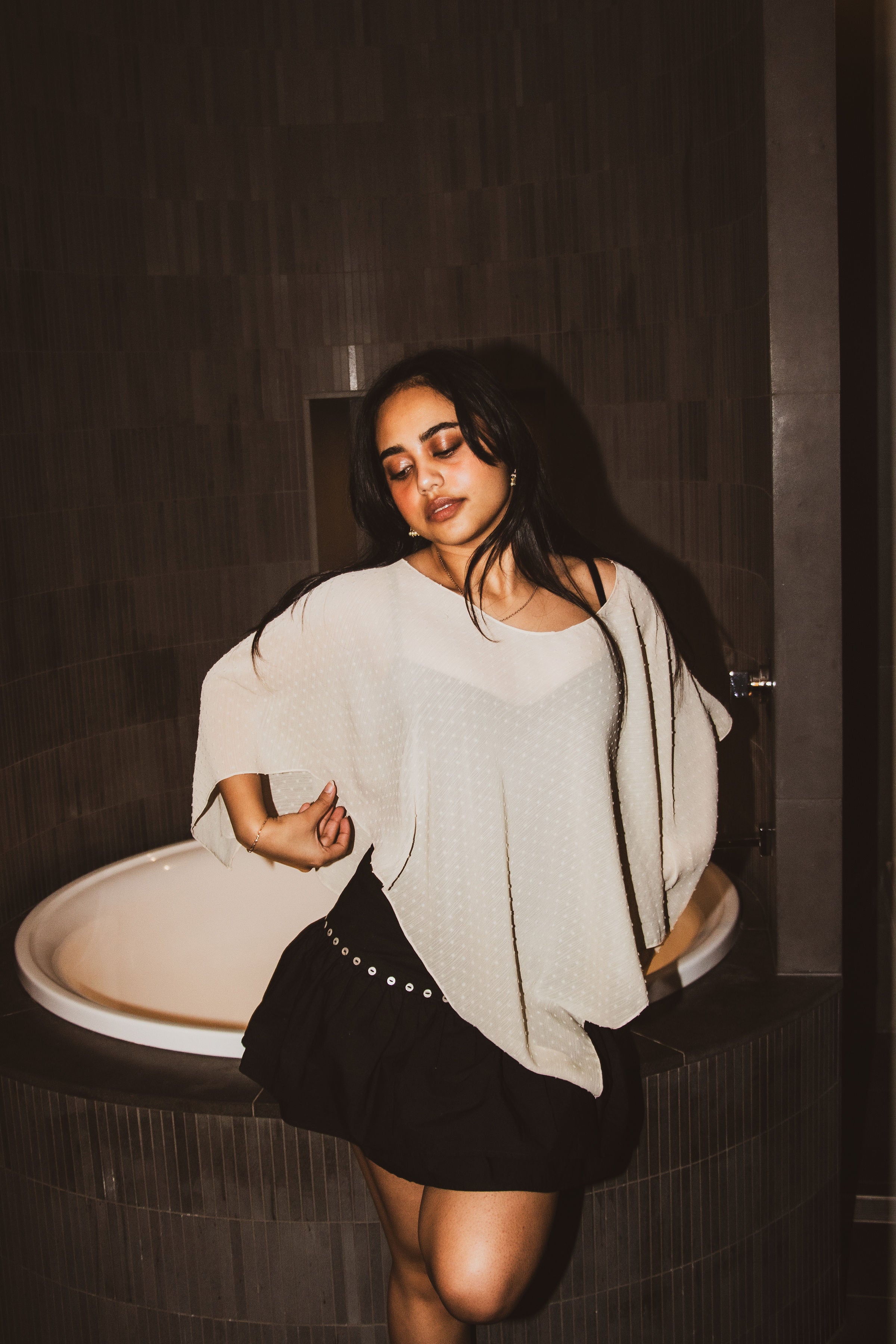 A woman posing in a bathroom next to a bathtub, wearing a loose, oversized light beige knit top and a black skirt with silver studs.