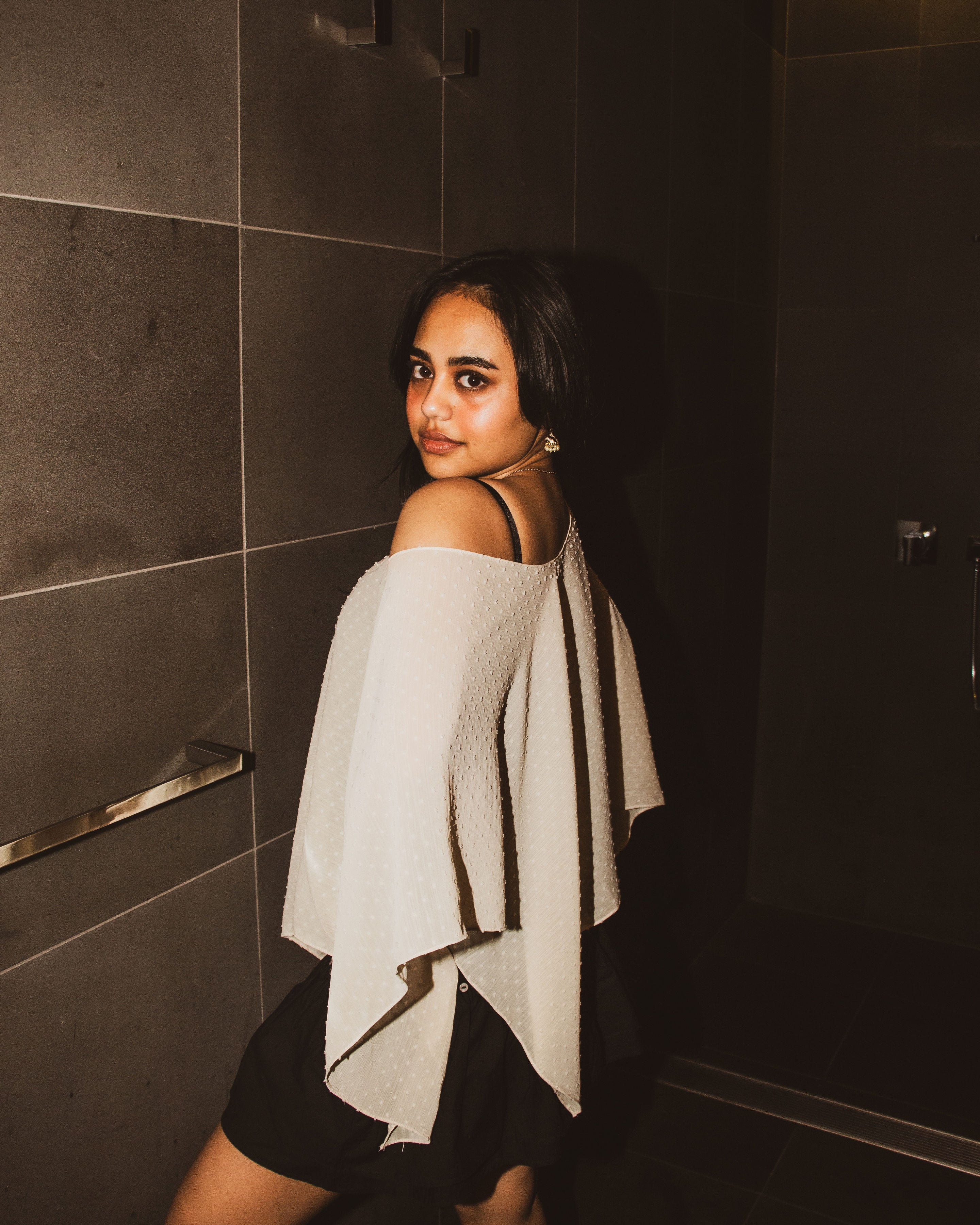 A woman in a black dress wearing a textured off-the-shoulder white cape draped over her shoulders, posing in a dark tiled bathroom.