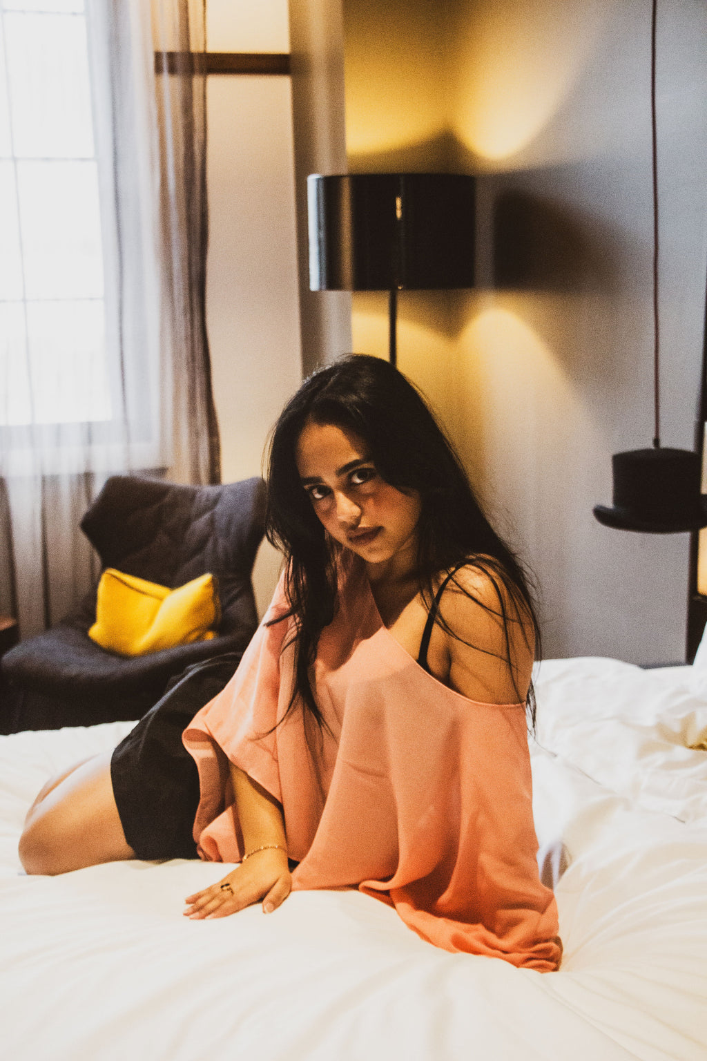A woman with long dark hair sits on a white bed in a bedroom, wearing a peach-pink off-the-shoulder knit top and a black skirt. A black floor lamp, a gray armchair with a yellow pillow, and a window with sheer curtains are visible in the background.