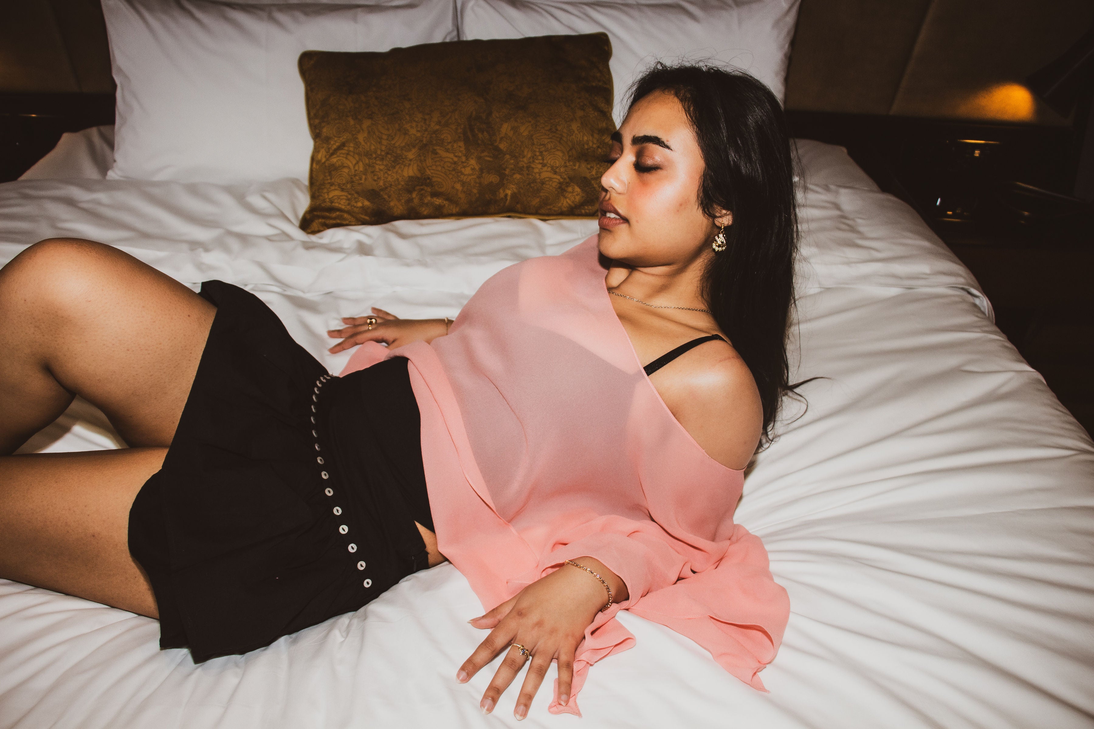 A woman reclining on a bed wearing a sheer light pink off-the-shoulder blouse and a black skirt with silver button details.