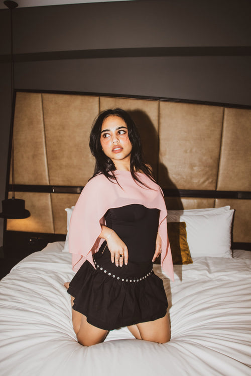 A woman models a black and pink dress while kneeling on a bed in a modern bedroom setting.
