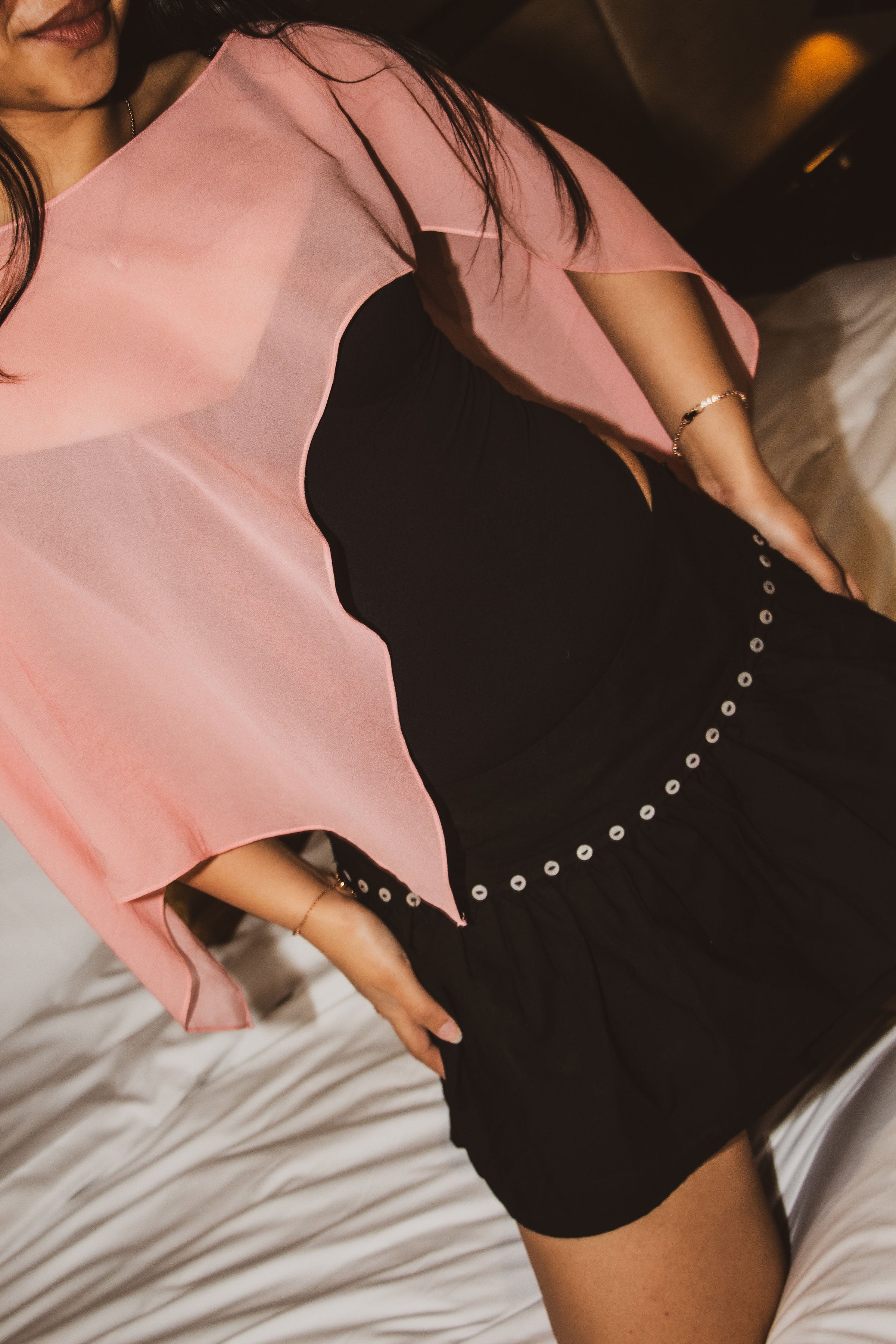 A woman wearing a black dress with a sheer pink overlay top, sitting on a bed with white sheets.